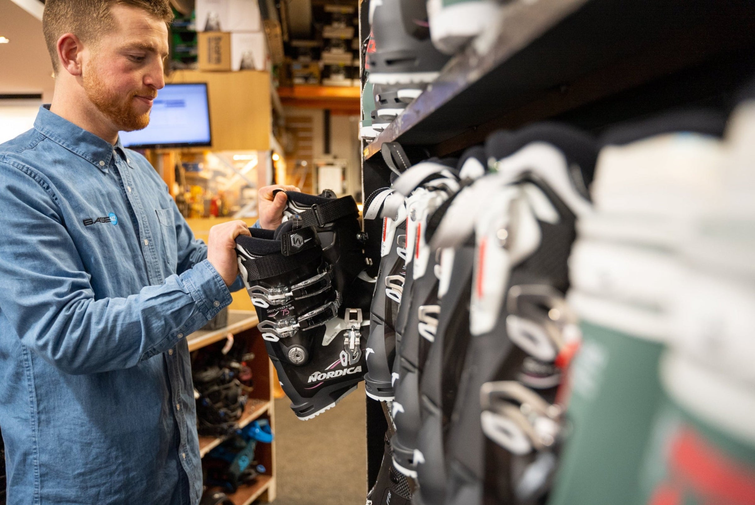 Man examining ski boots in a store with shelves and equipment in the background